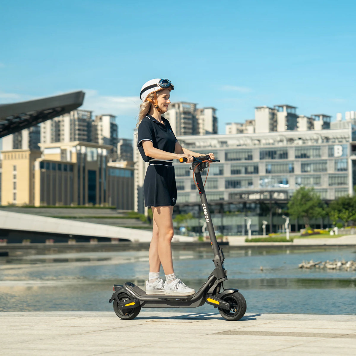 Woman wearing helmet riding a black Cunfon RS500 electric scooter in the city with a lake and buildings in the background.