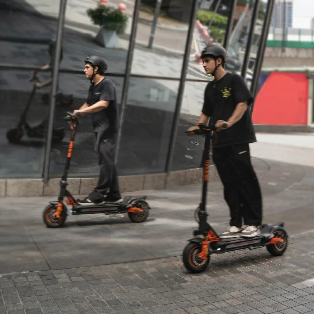 Rider wearing a black safety helmet riding a black and orange electric scooter on a city sidewalk next to a glass building reflection.