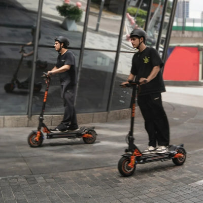 Rider wearing a black safety helmet riding a black and orange electric scooter on a city sidewalk next to a glass building reflection.